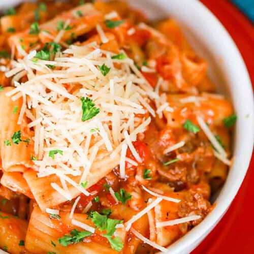 A bowl of rigatoni pasta topped with tomato sauce, Italian sausage, grated cheese, and garnished with chopped parsley. The delicious creation resembles a crock pot rigatoni recipe and sits in a white bowl against a surface with colorful background accents.