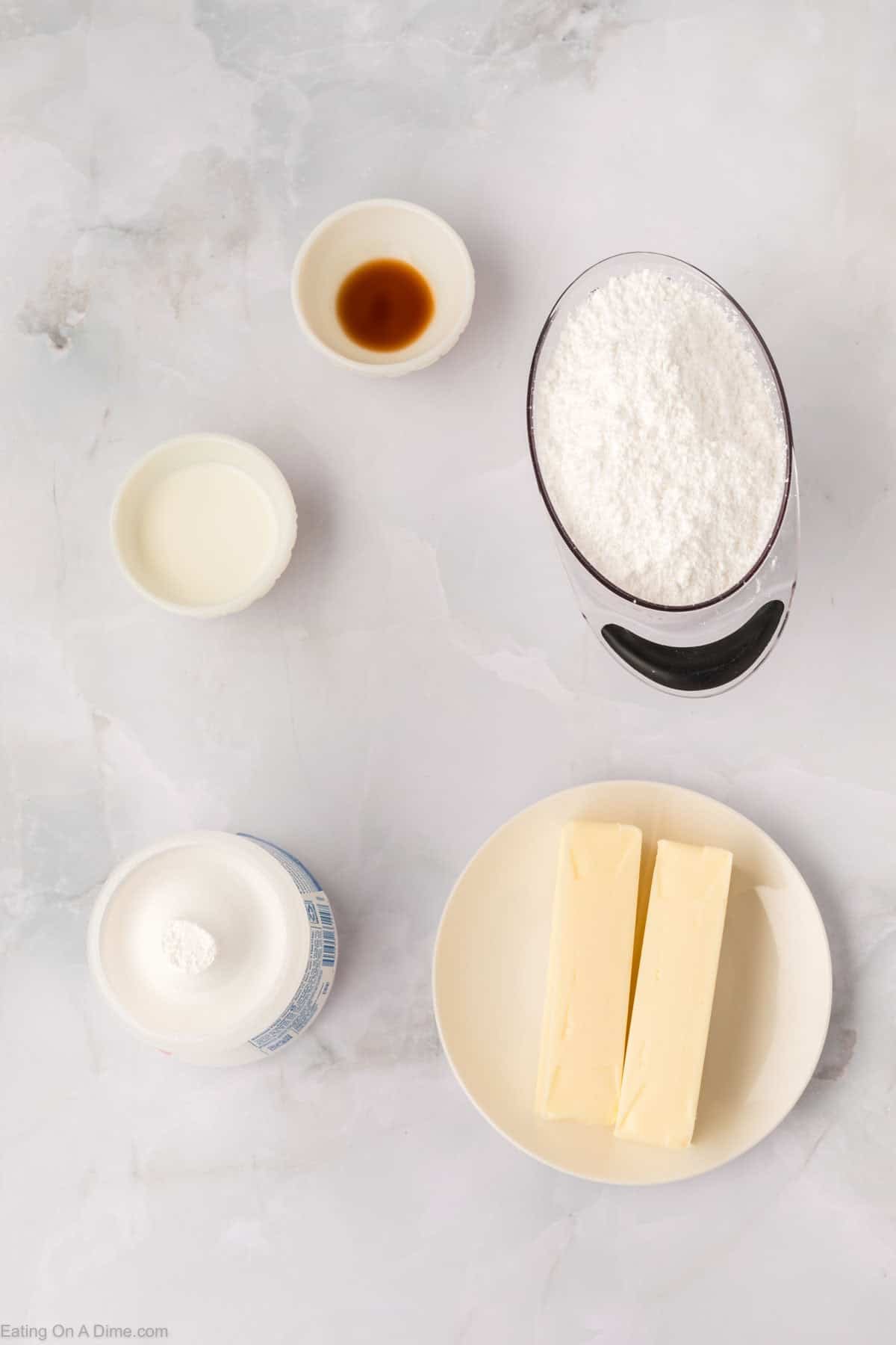 Ingredients for Marshmallow Fluff Frosting Recipe on a marble surface: a cup of powdered sugar, two sticks of butter on a plate, a small bowl of vanilla extract, a small bowl of milk, and an open container of shortening with a tablespoon beside.