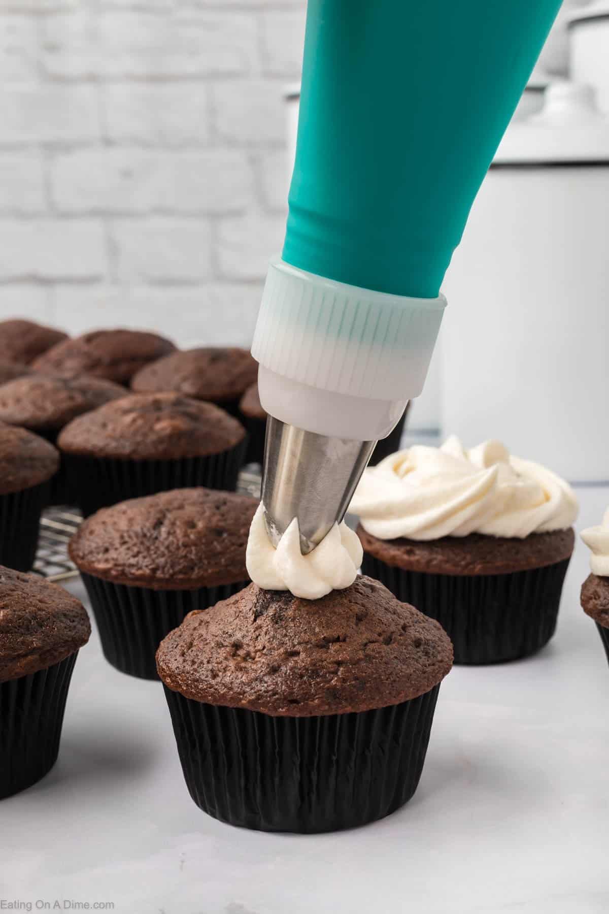 A close-up of a hand holding a piping bag with Marshmallow Fluff frosting, artfully decorating a chocolate cupcake. Other chocolate cupcakes linger in the background, framed by a brick wall and kitchen canisters.