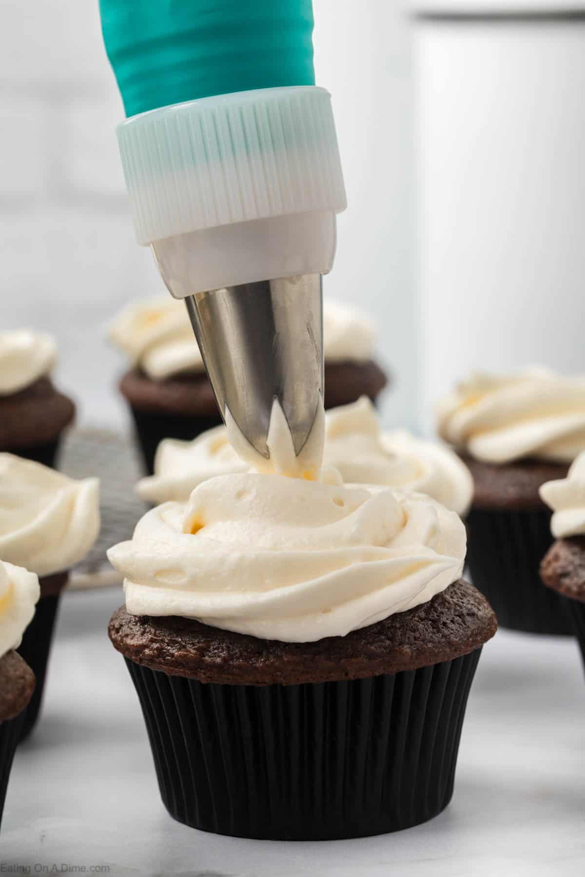 A close-up of a chocolate cupcake being frosted with vanilla icing from a piping bag showcases the technique behind marshmallow fluff frosting. Other frosted cupcakes rest on a cooling rack, completing the sweet scene set in a bright kitchen.