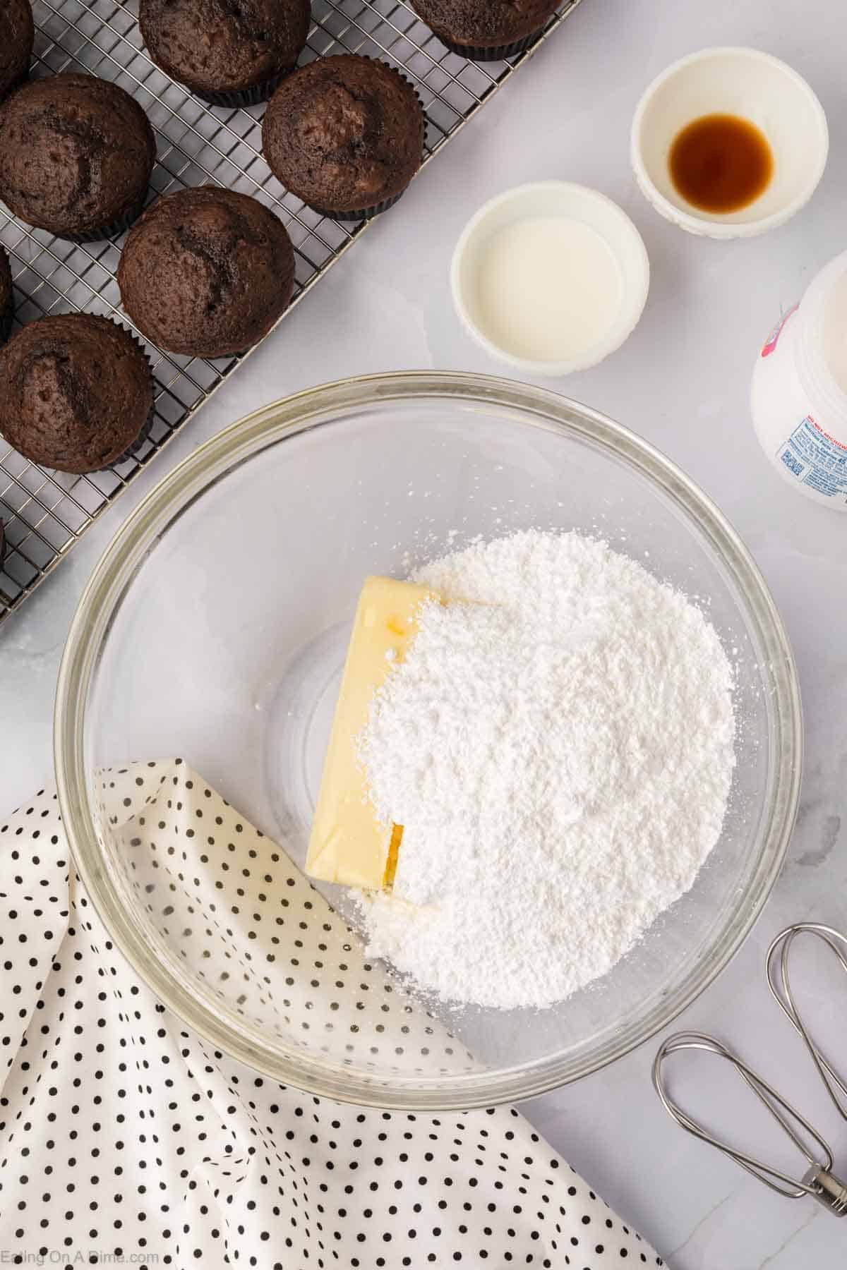 A mixing bowl with butter and powdered sugar awaits transformation into a Marshmallow Fluff frosting recipe, surrounded by small bowls of cream and vanilla extract. A polka-dotted cloth and whisk are nearby, while chocolate muffins cool on a wire rack in the background.