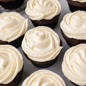 Chocolate cupcakes with creamy swirls of marshmallow fluff frosting are arranged closely together. Each cupcake is in a dark wrapper, placed on a light surface. A cooling rack is partially visible in the background.