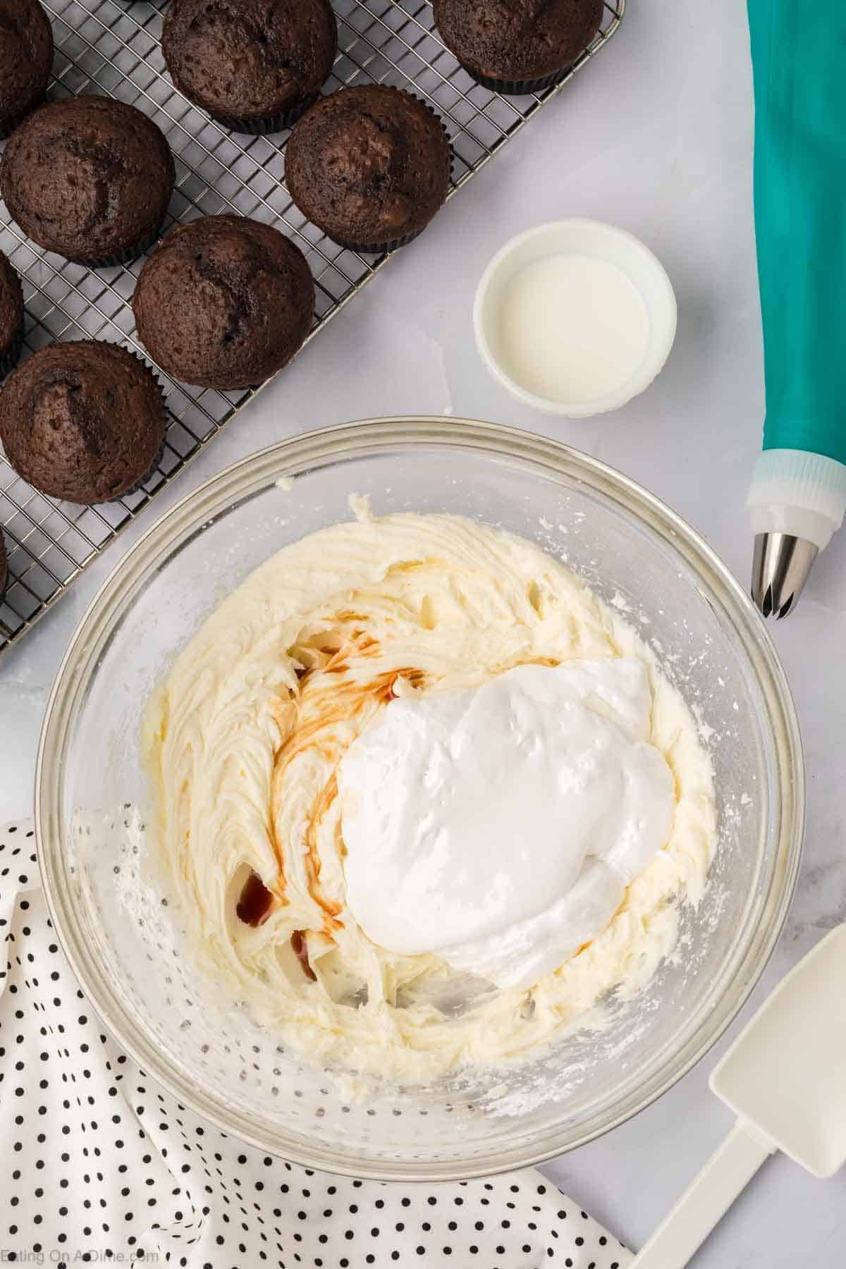 A glass bowl brims with creamy Marshmallow Fluff frosting, a swirl of whipped topping, and a splash of vanilla. Nearby are chocolate muffins on a cooling rack, a small bowl of milk, a teal piping bag, and a polka-dot cloth.