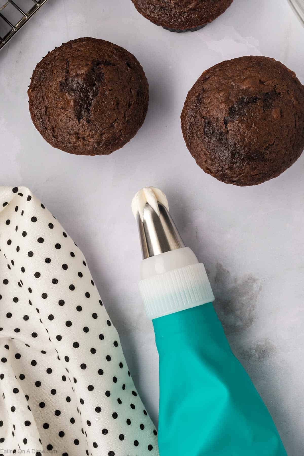 Three chocolate muffins sit on a light surface beside a teal piping bag fitted with a metal nozzle, ready for you to try your hand at creating Marshmallow Fluff Frosting. Nearby, a white cloth adorned with black polka dots adds an extra touch of charm to the scene.