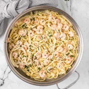 A large stainless steel pot filled with creamy Shrimp Alfredo fettuccine pasta garnished with chopped parsley. A grey and white striped cloth is draped on the left side of the pot, and a marble surface is visible underneath.