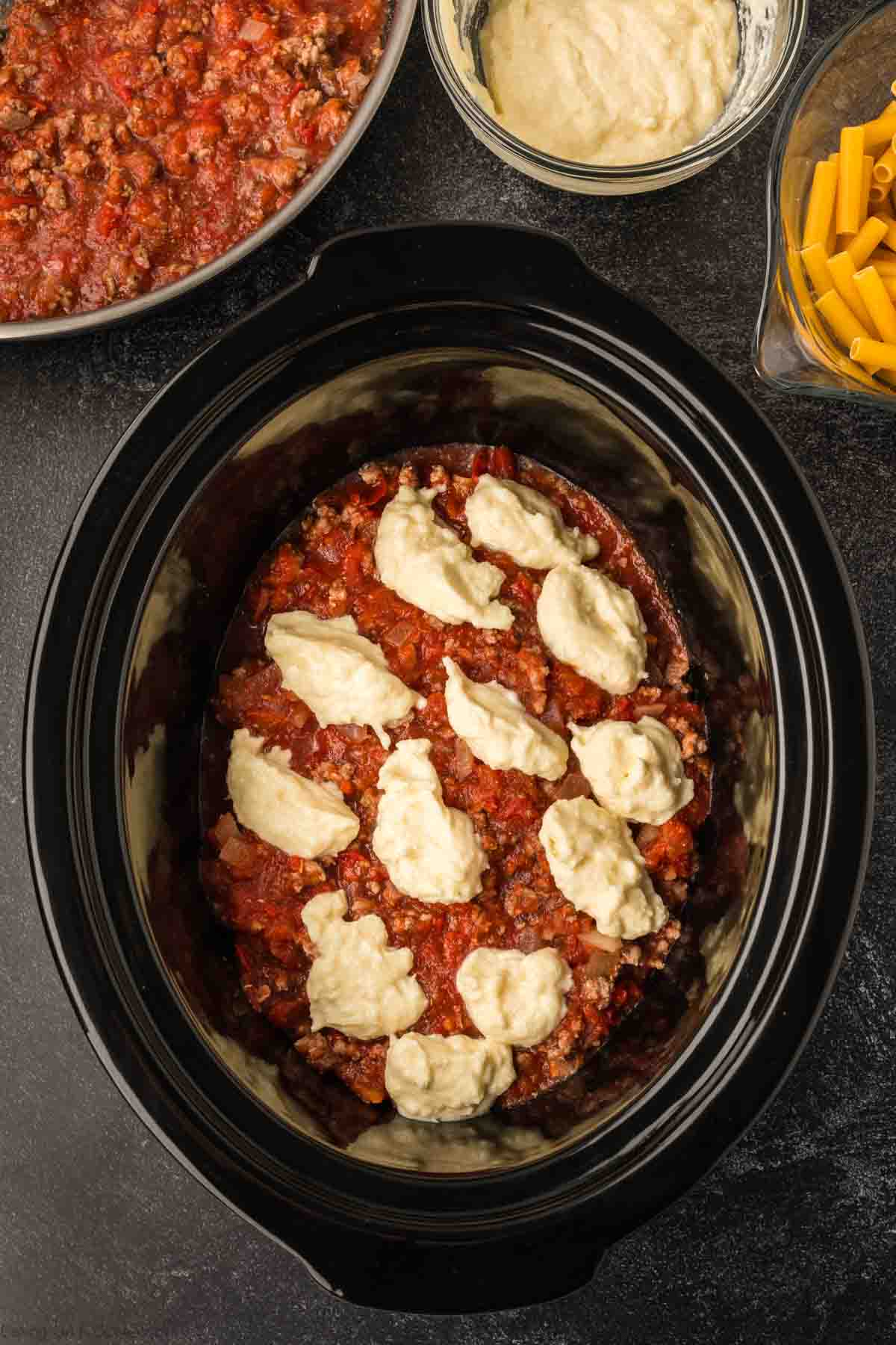 A slow cooker is filled with layers of tomato sauce, ground meat, and dollops of ricotta cheese, crafting the perfect baked ziti recipe. Nearby are bowls of more sauce, ricotta, and uncooked pasta on a dark surface.