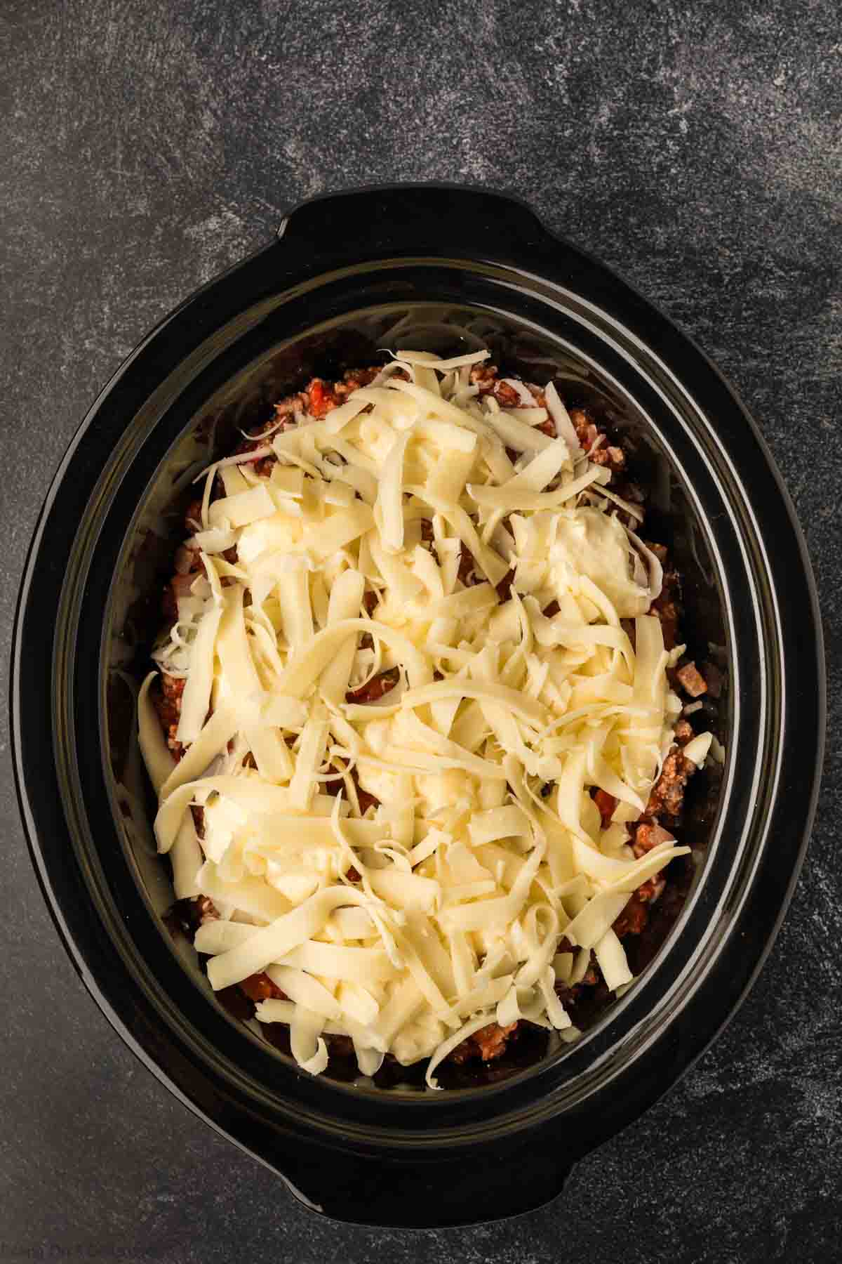 A slow cooker showcases a delightful baked ziti recipe, with grated cheese melting over layers of cooked ground meat and vegetables, set against a dark textured background.