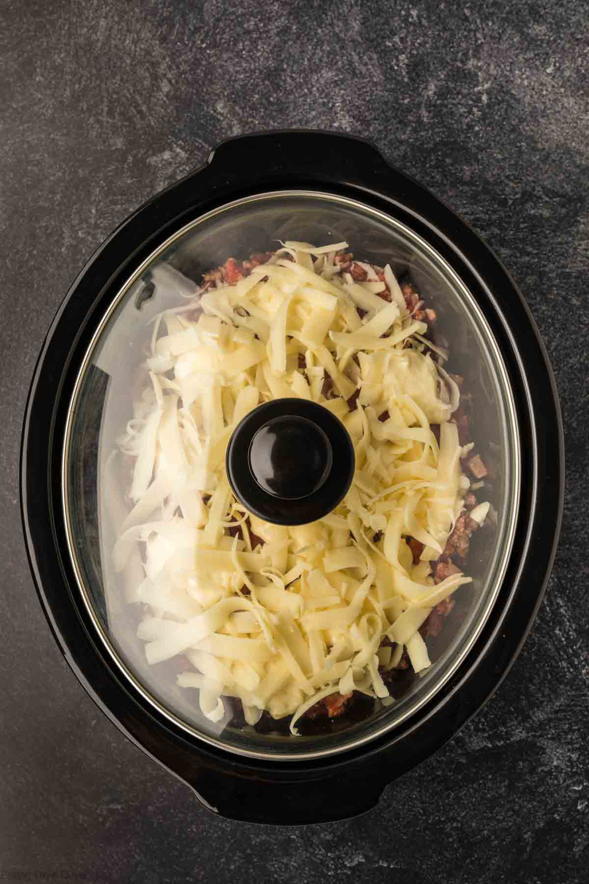 A slow cooker filled with savory baked ziti, topped with melted shredded cheese, is visible through the glass lid. The dish sits against a dark, textured background, highlighting the delicious simplicity of this comforting recipe.