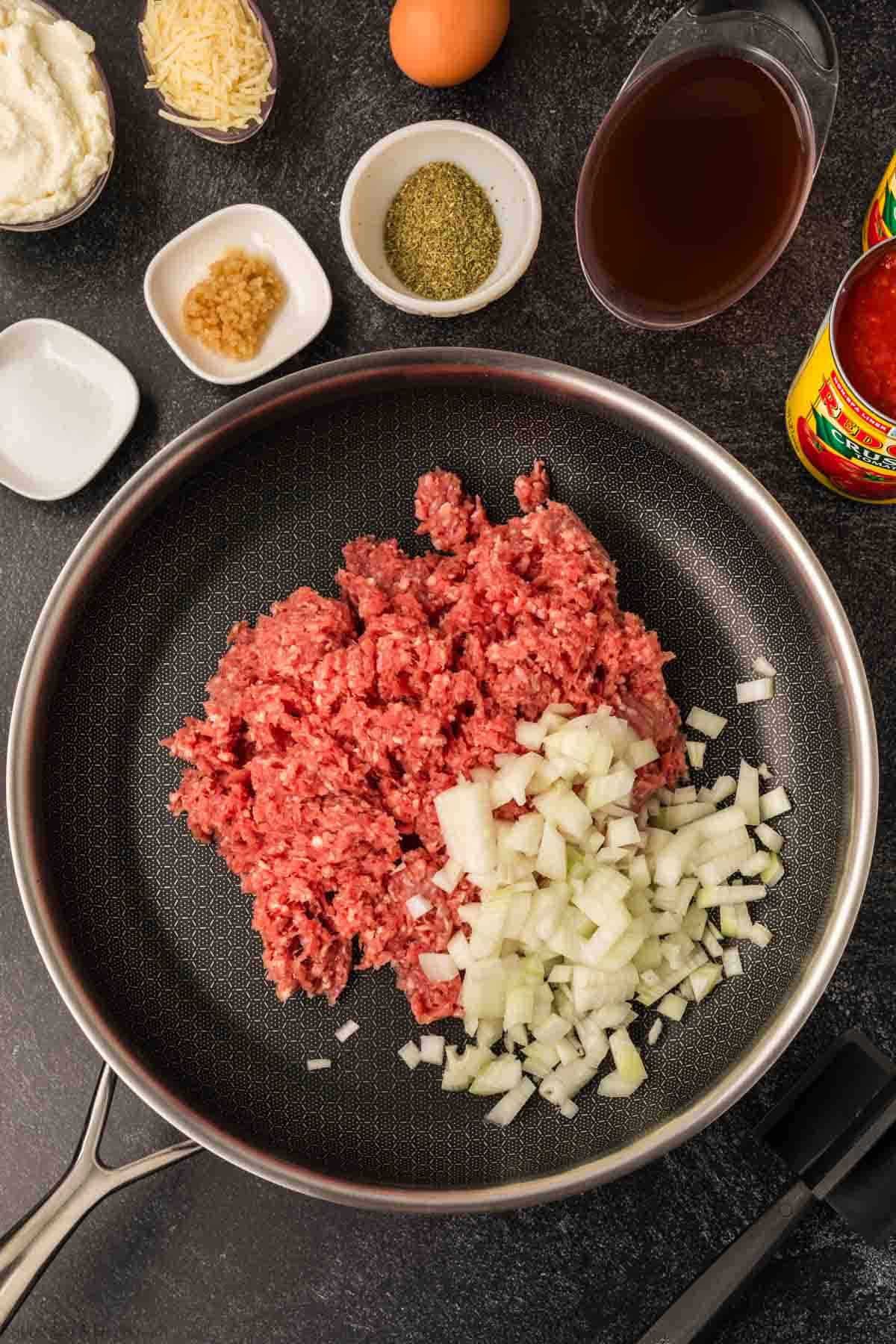 A frying pan on a dark surface holds ground meat and chopped onions, perfect for starting a slow cooker baked ziti recipe. Surrounding the pan are ingredients like cheese, an egg, minced garlic, herbs, broth, and a can of tomatoes—all poised to create a hearty dish.