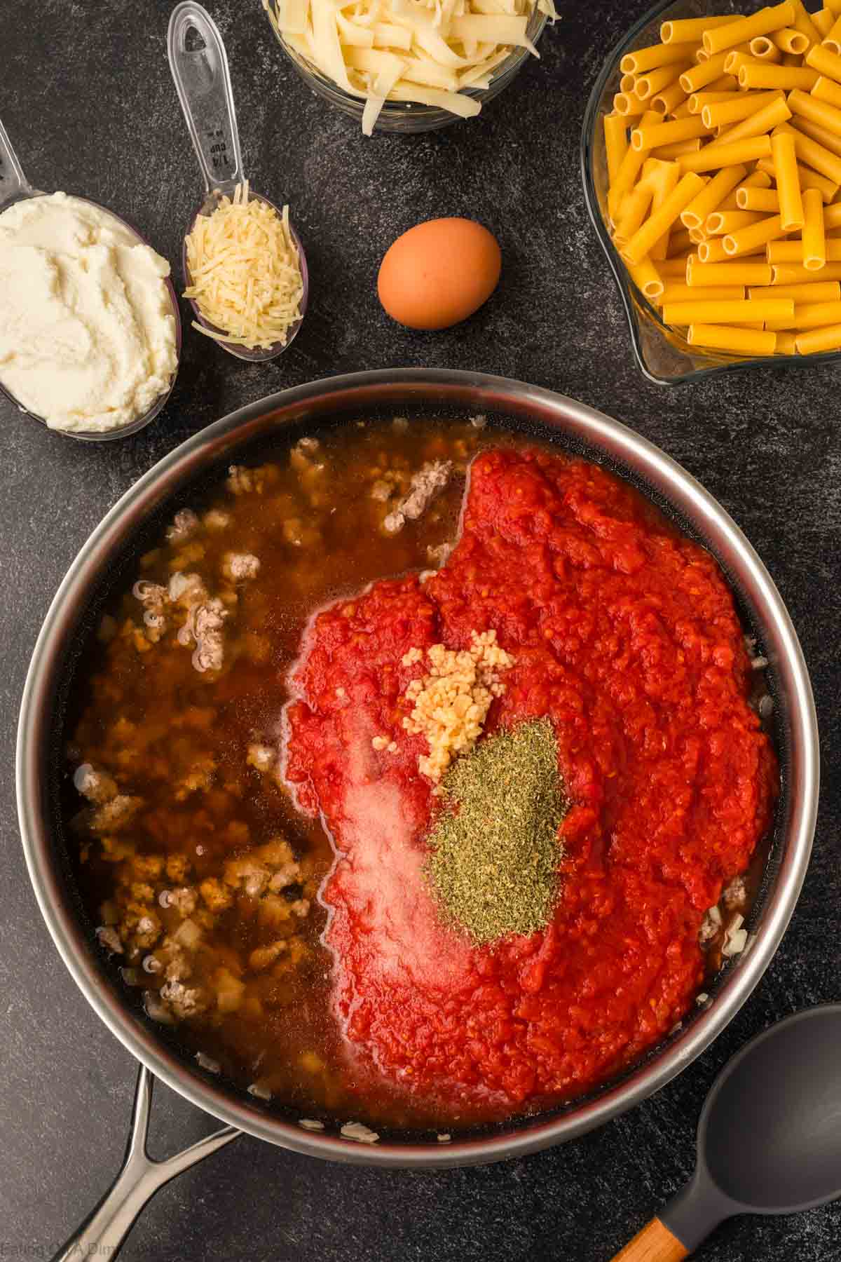 A skillet with tomato sauce, minced garlic, and herbs is being prepared for a Slow Cooker Baked Ziti Recipe. Surrounding it are ingredients: dry ziti pasta, an egg, ricotta, grated cheese, and shredded mozzarella on a dark countertop.