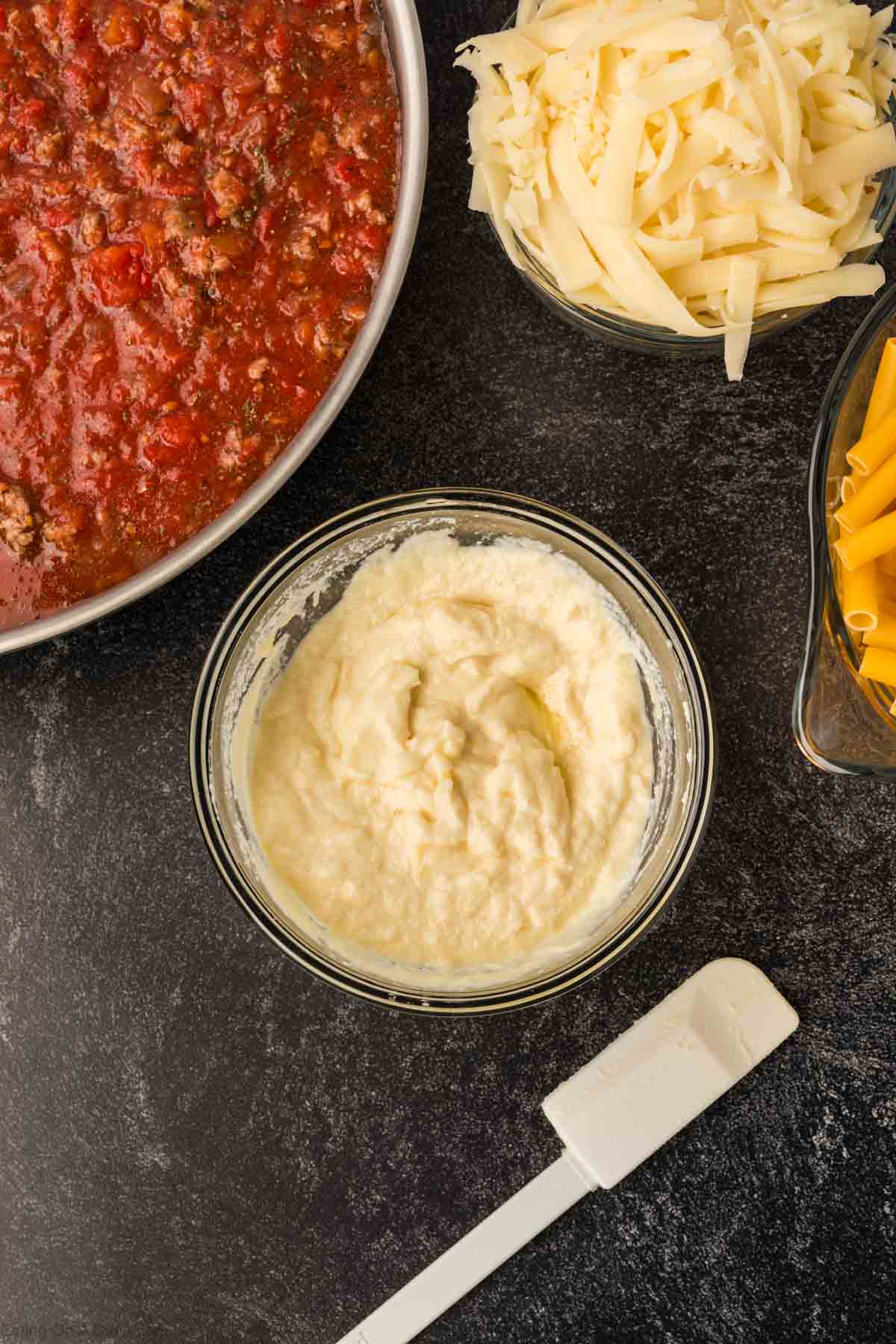 Overhead view of lasagna ingredients on a dark surface: a bowl of red meat sauce reminiscent of slow cooker baked ziti, creamy ricotta cheese mixture, shredded cheese, uncooked lasagna noodles, and a white spatula.