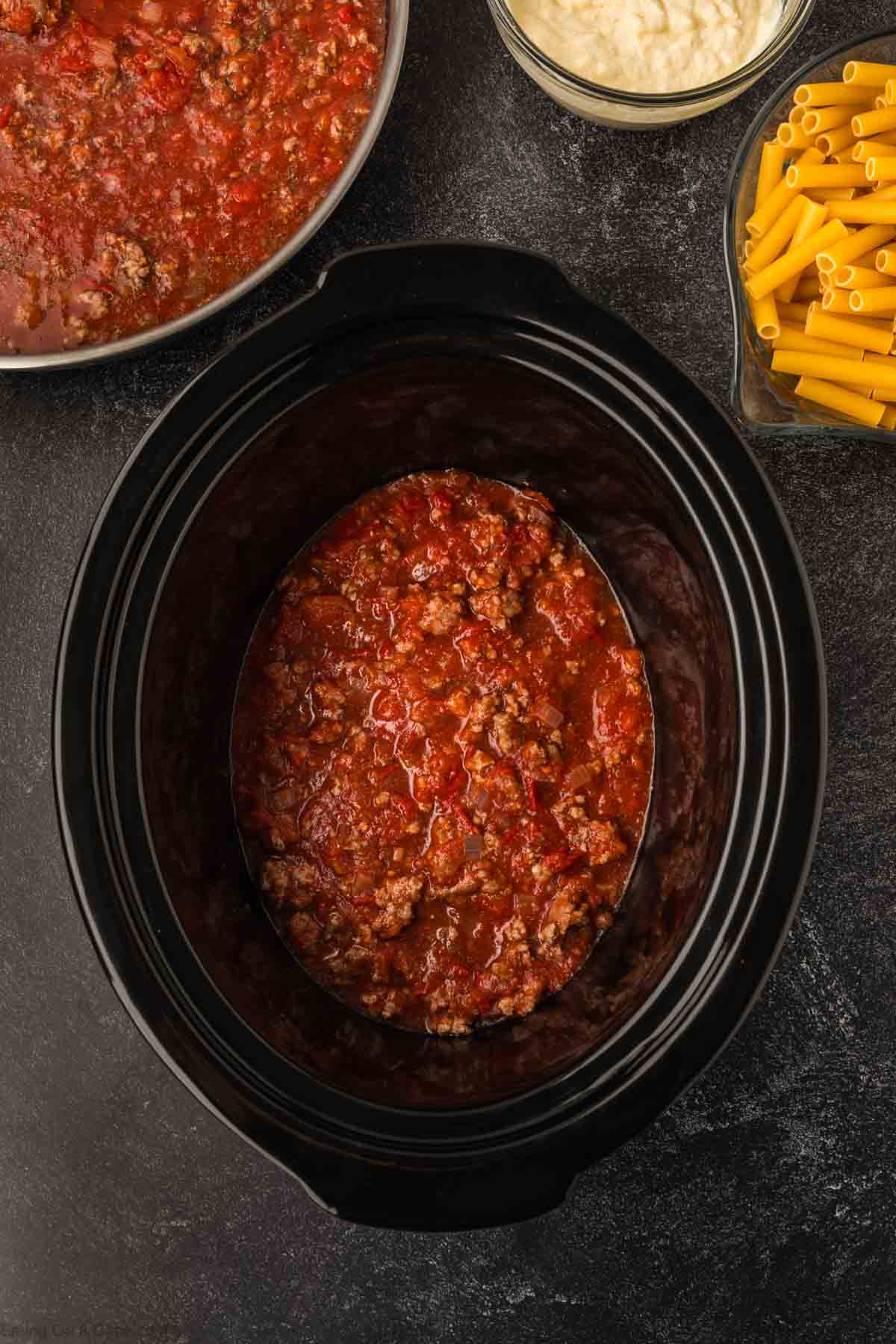 Top view of a black slow cooker filled with a tomato-based meat sauce, perfect for crafting a Slow Cooker Baked Ziti Recipe. Surrounding it are a bowl of cooked sauce, uncooked penne pasta, and grated cheese, all displayed on a dark surface.