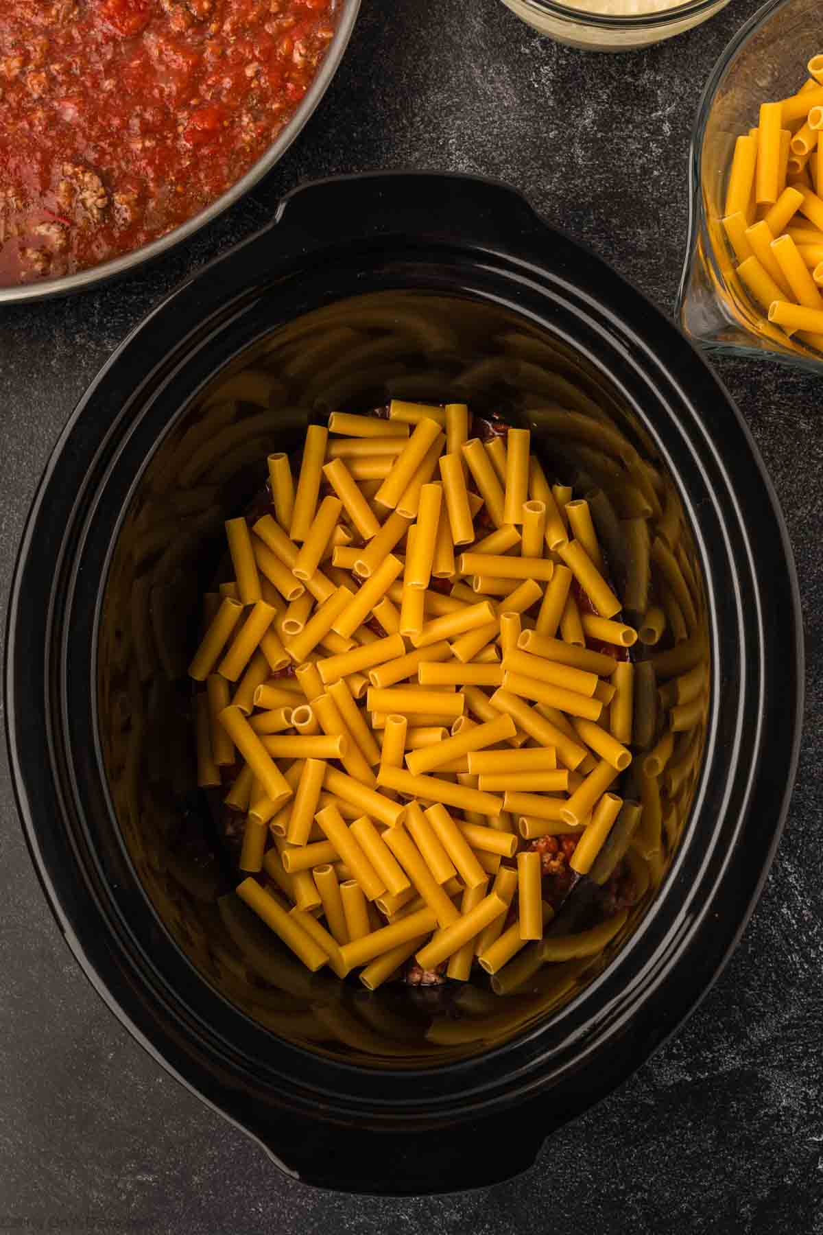 A top-down view of a black slow cooker showcasing an uncooked Slow Cooker Baked Ziti recipe. To the side, a pan with vibrant red sauce sits alongside bowls of extra pasta and freshly grated cheese on a dark surface.