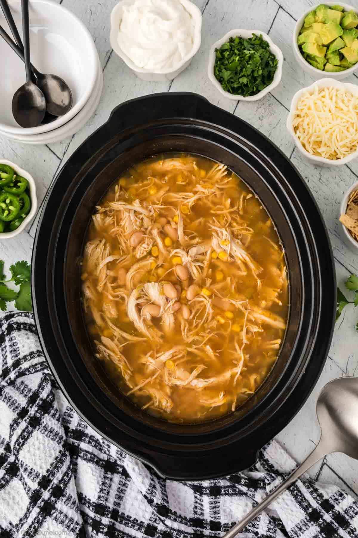 A black slow cooker brims with Slow Cooker White Chicken Chili, featuring shredded chicken, corn, and beans. Surrounding bowls boast toppings like cheese, sour cream, jalapeños, avocado, and cilantro. A black and white checkered towel and two spoons await nearby.