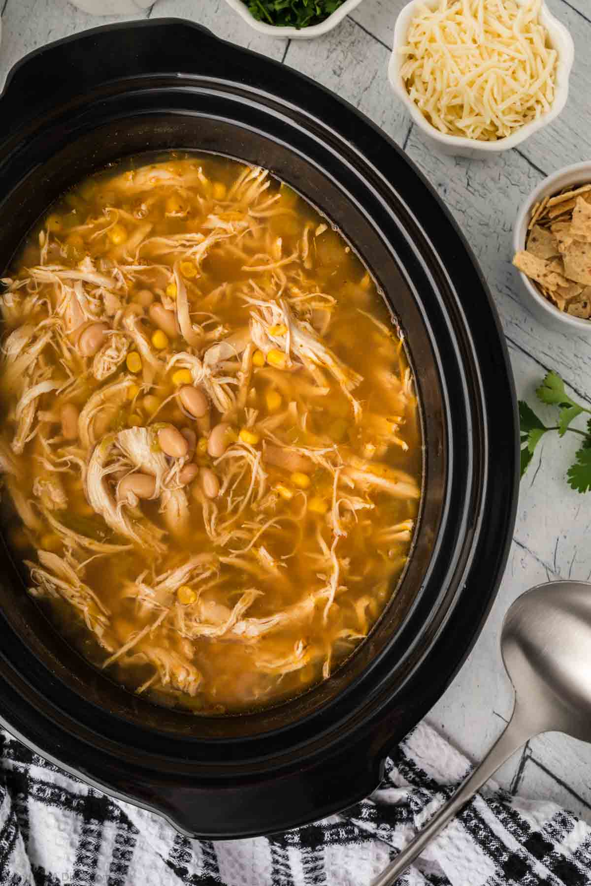 A slow cooker filled with Slow Cooker White Chicken Chili combines shredded chicken, beans, corn, and broth. Surrounding the cooker are small bowls with shredded cheese, tortilla chips, and cilantro. A silver spoon and checkered cloth are nearby, completing the cozy scene.