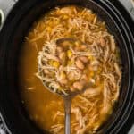 A close-up of a slow cooker filled with white chicken chili showcases shredded chicken, beans, and corn. A ladle lifts some soup, revealing a medley of ingredients in the light, flavorful broth.