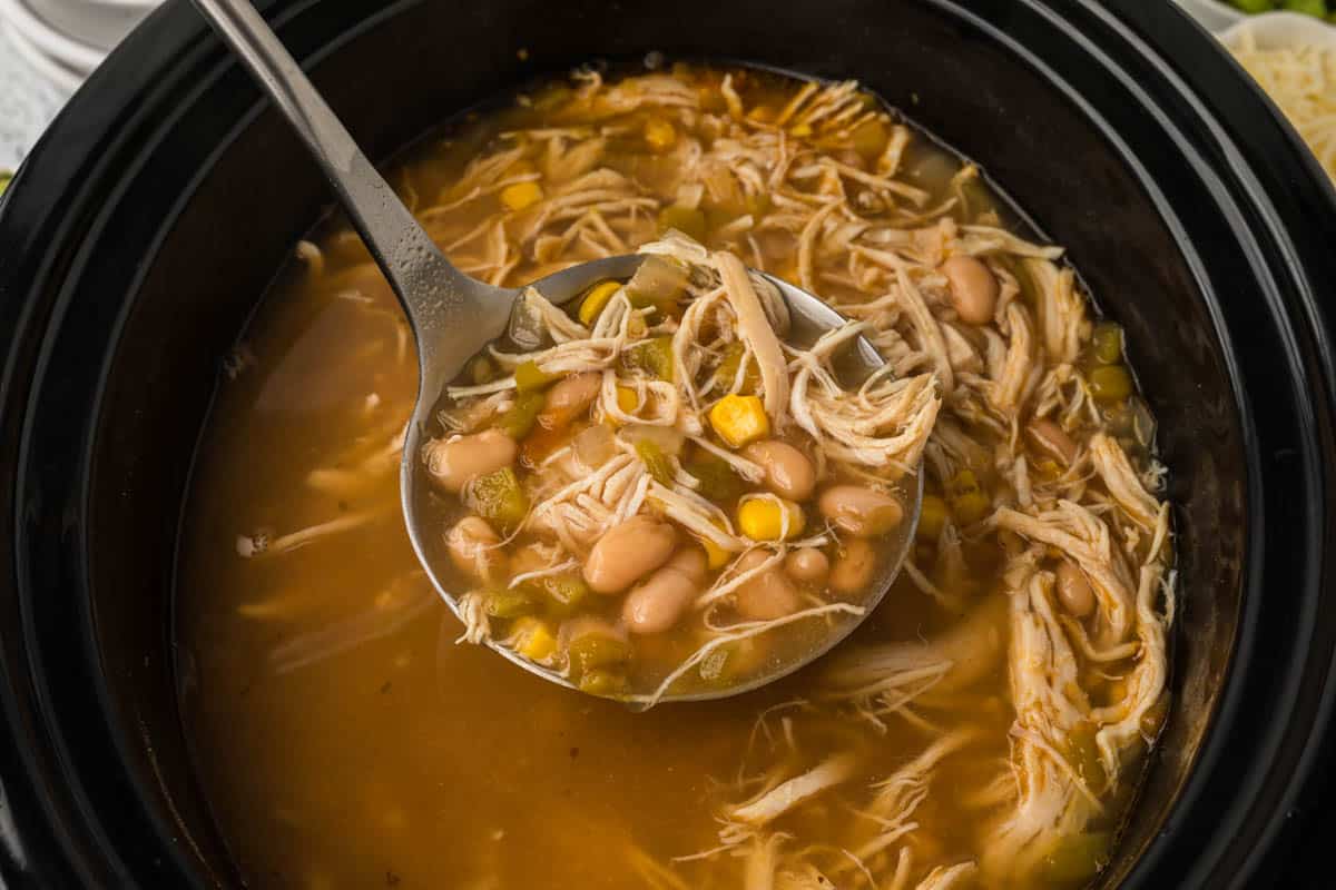 A ladle scoops up a serving of Slow Cooker White Chicken Chili from a black slow cooker, brimming with shredded chicken, white beans, corn, and green peppers in a savory broth.