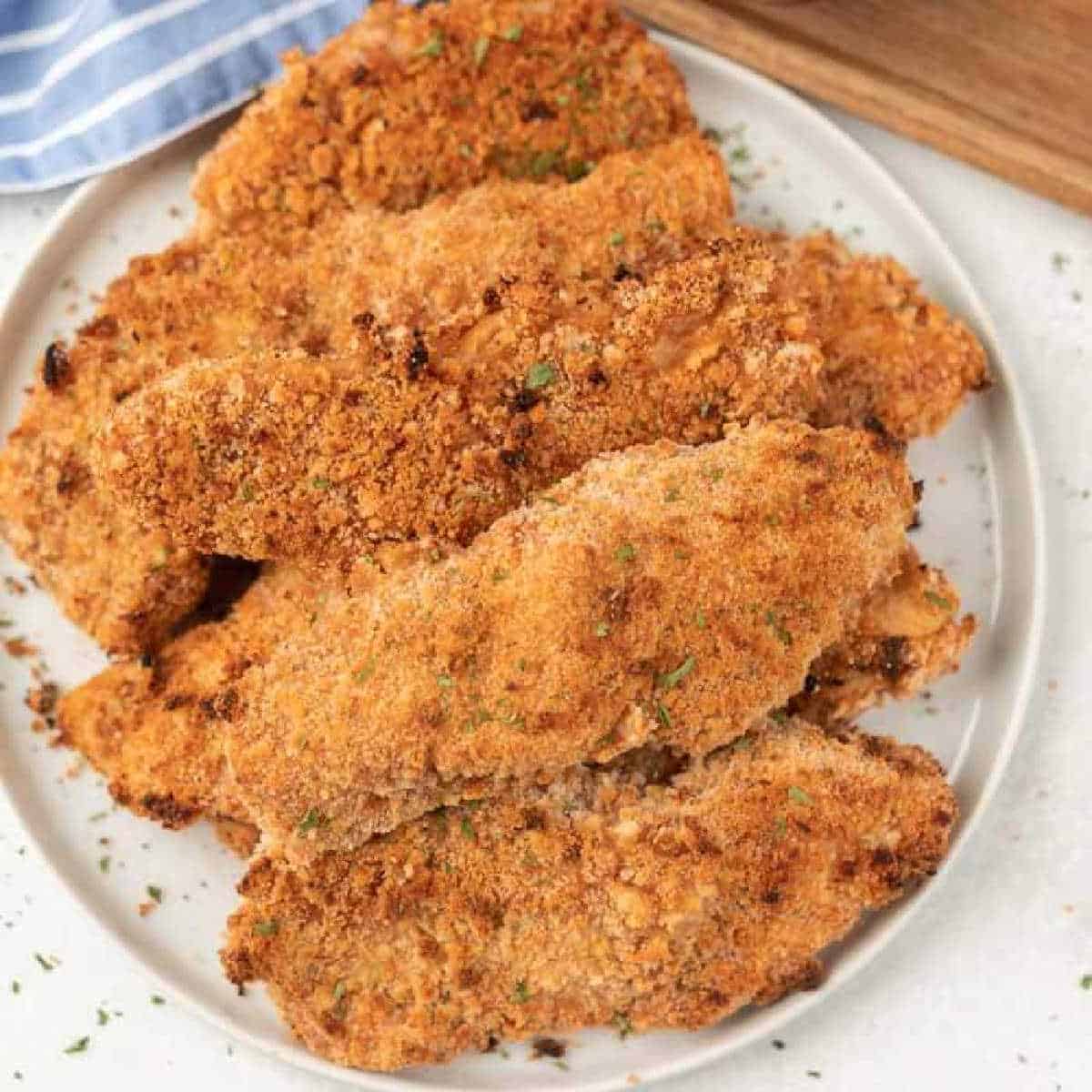 A plate piled with crispy, golden-brown oven-baked breaded chicken tenders, garnished with a sprinkle of herbs, is displayed. The tenders appear freshly cooked and are arranged neatly on a white round plate. A striped cloth and wooden board are partially visible.