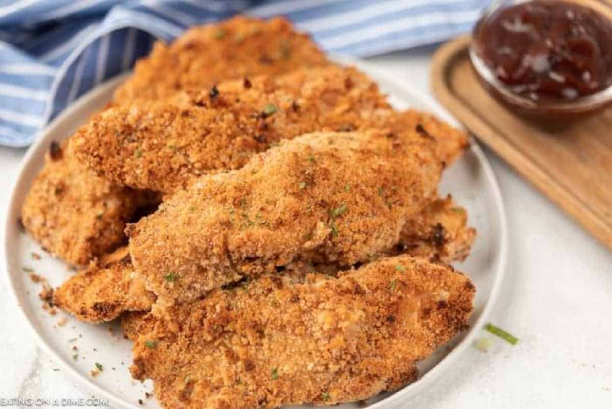 A plate of golden-brown, oven-baked breaded chicken tenders garnished with herbs. A small bowl of dipping sauce sits in the background, beside a blue and white striped cloth.