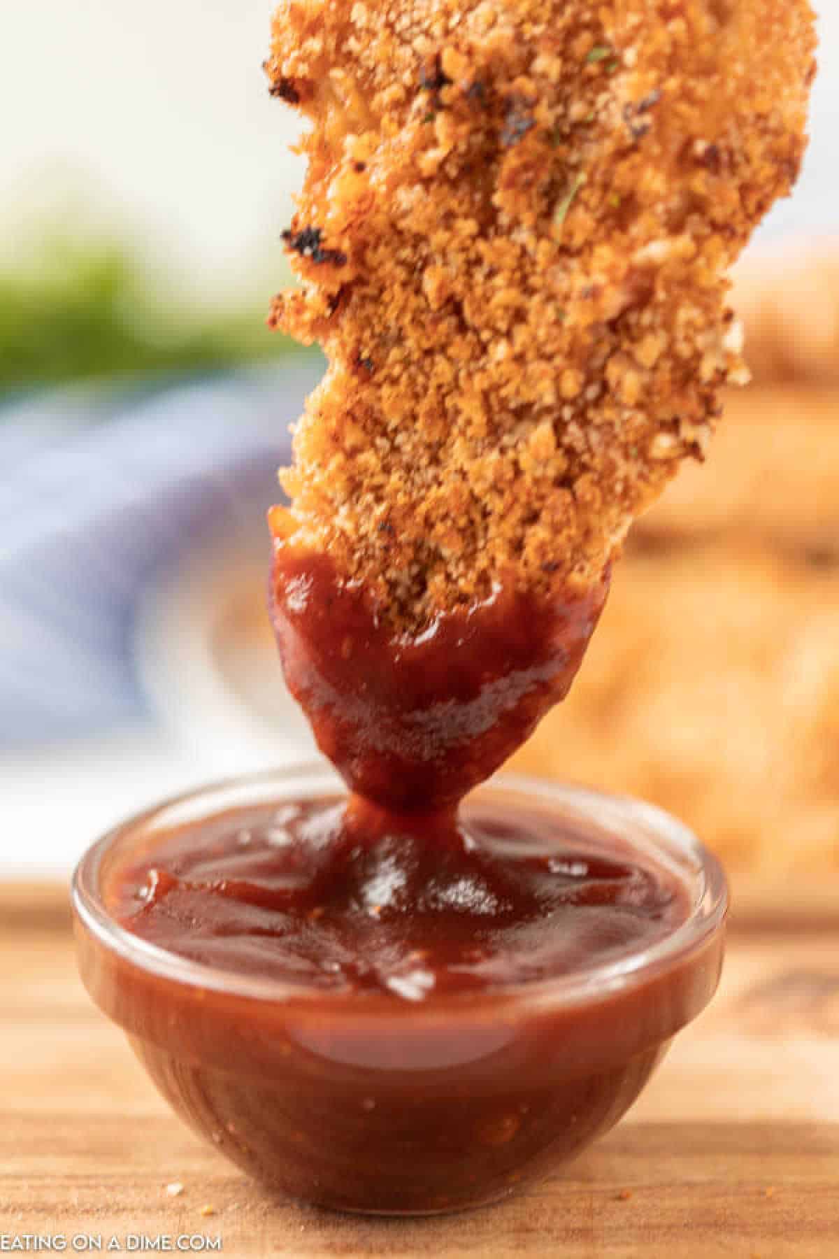 Close-up of crispy oven baked breaded chicken tenders being dipped into a small glass bowl of rich, red barbecue sauce. The background is blurred, focusing attention on the dipping motion and texture of the food.