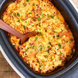 A crock pot beef noodle casserole brimming with cooked noodles, melted cheese, and ground meat is garnished with chopped parsley. A wooden spoon rests inside, ready to serve from the black rectangular pan on a wooden table.