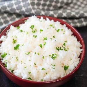 A red bowl brimming with fluffy Instant Pot white rice, elegantly garnished with chopped parsley. In the background, a patterned gray and white cloth adds a touch of subtle style.