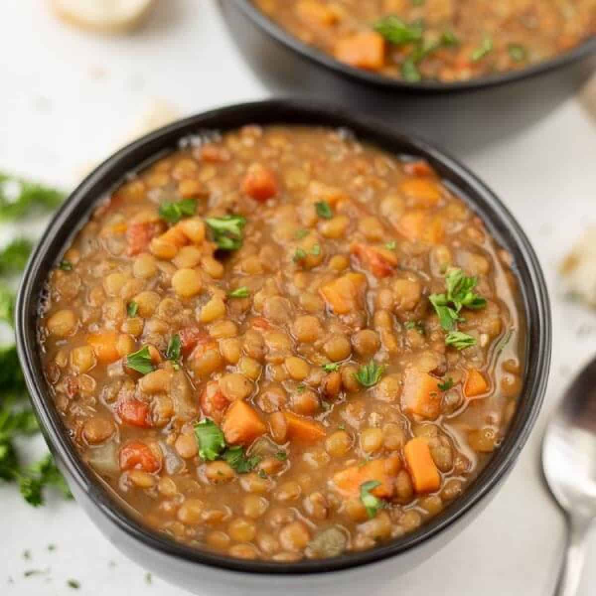 A bowl of Instant Pot lentil soup filled with tender lentils, diced carrots, and garnished with fresh parsley. Another bowl of this delicious recipe is partially visible in the background. A spoon rests beside the bowl on a pristine white surface.