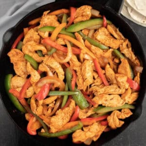 A cast iron skillet filled with sizzling Skillet Chicken Fajitas showcases strips of chicken, red and green bell peppers, and onions all cooked to golden brown perfection. Warm tortillas await in the background, ready to wrap up this savory delight.