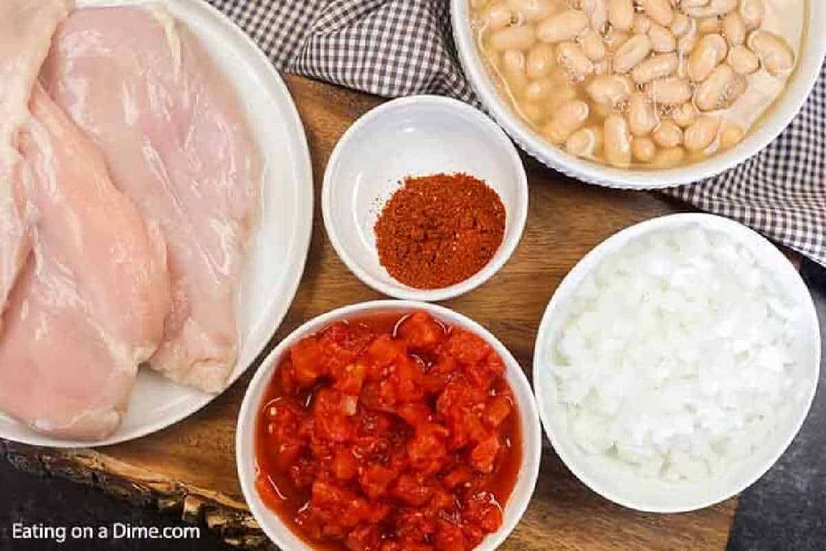 Ingredients for a delightful Slow Cooker White Chicken Chili are laid out on a wooden surface: raw chicken breasts rest on a white plate, beside bowls of chopped tomatoes, chili powder, white beans, and onions. A checkered cloth adds charm to this inviting culinary scene.