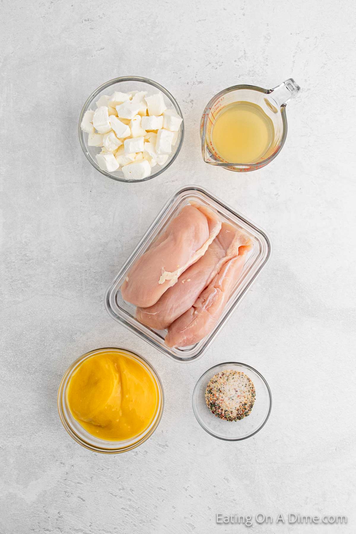 Top-down view of ingredients for a Slow Cooker Italian Chicken recipe on a light surface: raw chicken breasts in a glass container, a bowl of cubed cream cheese, a small jug of broth, a bowl of mustard, and mixed spices ready to create an aromatic delight.