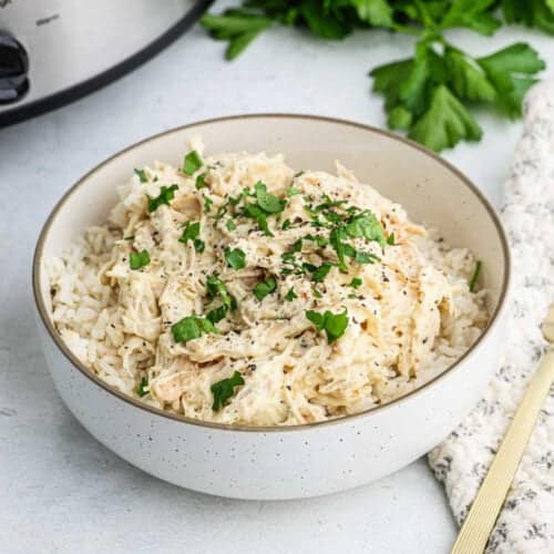 A bowl of creamy shredded chicken over rice, garnished with chopped parsley. This Slow Cooker Italian Chicken recipe comes to life against a backdrop of fresh parsley leaves and a textured cloth, promising a delightful meal that's both flavorful and effortless to prepare.