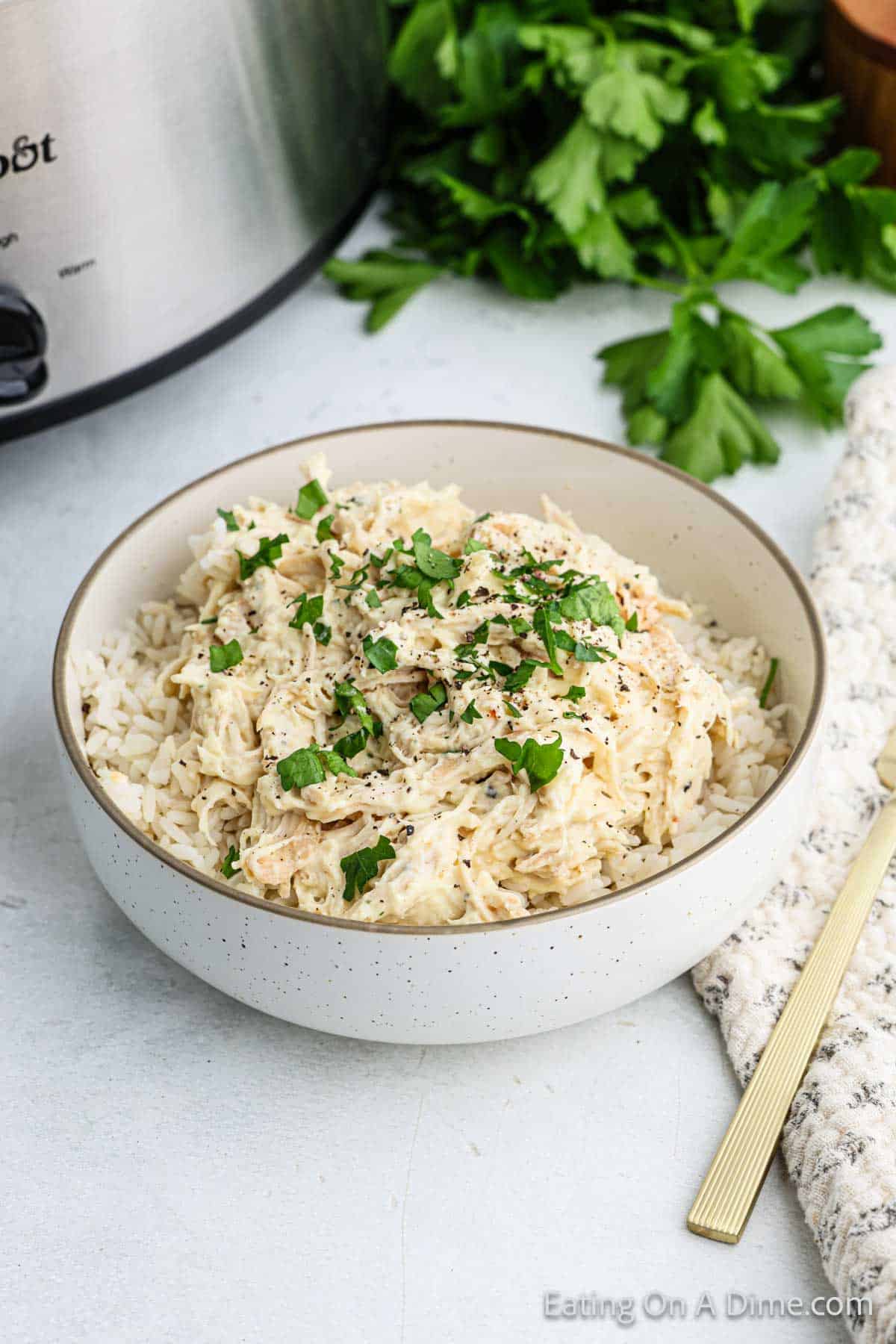 A bowl of creamy shredded Italian chicken over rice, garnished with chopped parsley. A gold fork is placed on the side, hinting at this easy slow cooker recipe, with fresh parsley and a slow cooker partially visible in the background.