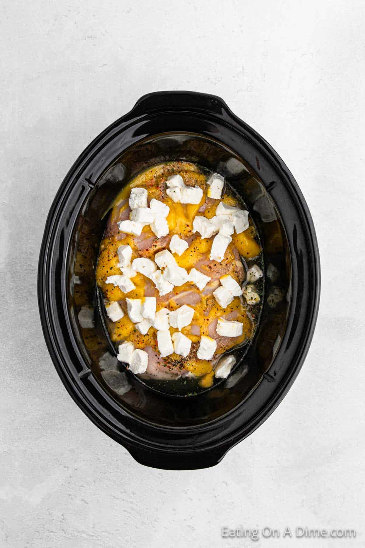 A black slow cooker brimming with ingredients for a delectable Italian chicken recipe, including chunks of cheese and visible herbs, ready to cook. The cooker sits elegantly on a textured light gray surface, promising a delightful culinary experience.