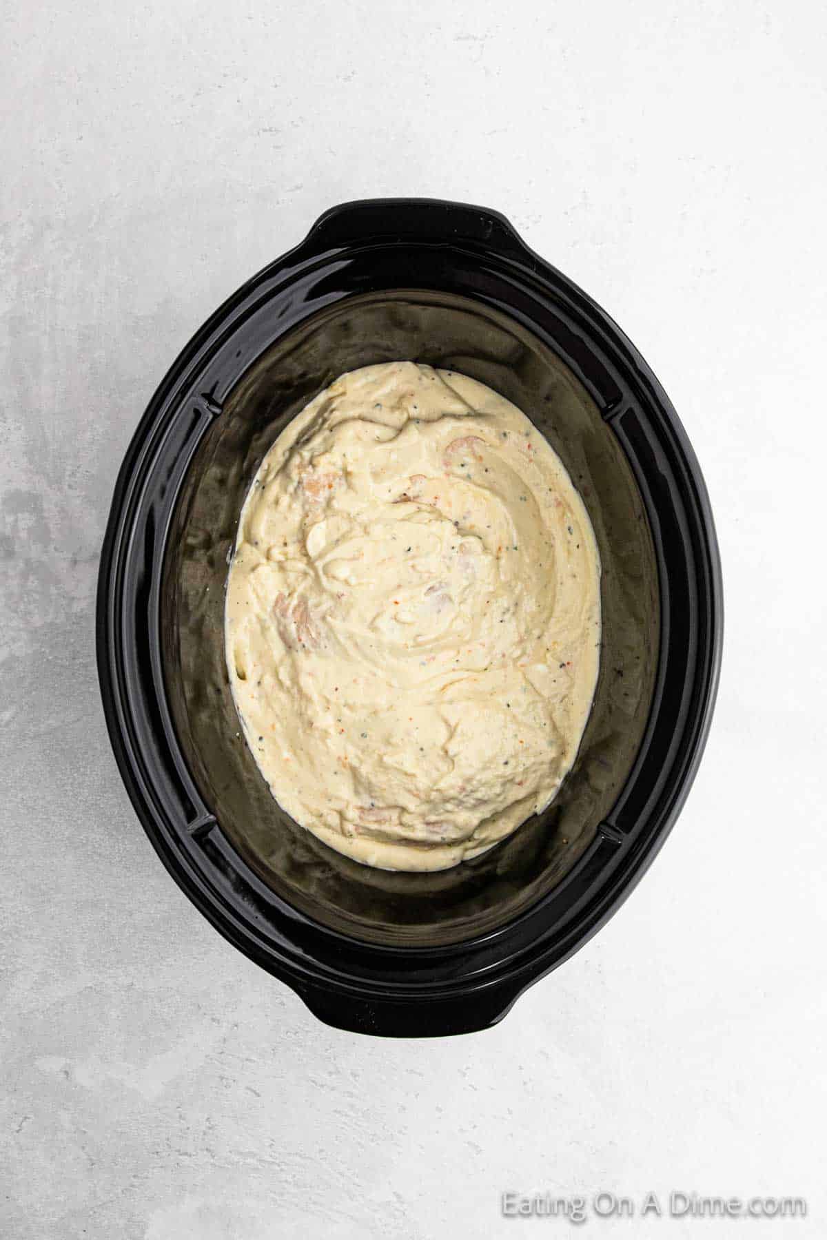 A slow cooker filled with a creamy, seasoned mixture, reminiscent of a savory Italian chicken recipe, sits on a gray textured surface. The dip, light in color with visible specks of seasoning, promises an inviting blend of flavors.