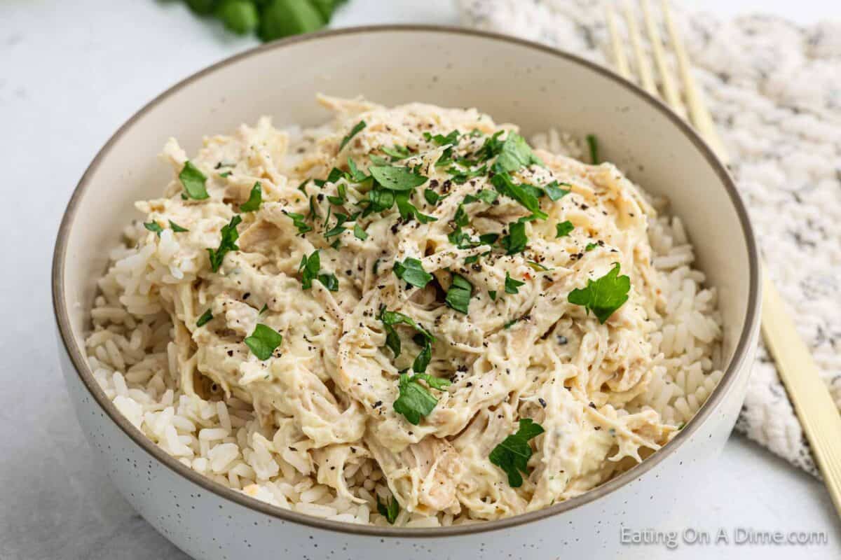 A bowl of shredded chicken in a creamy sauce, reminiscent of a Slow Cooker Italian Chicken Recipe, served over white rice and garnished with freshly chopped parsley. A fork and a textured napkin are placed beside the bowl.