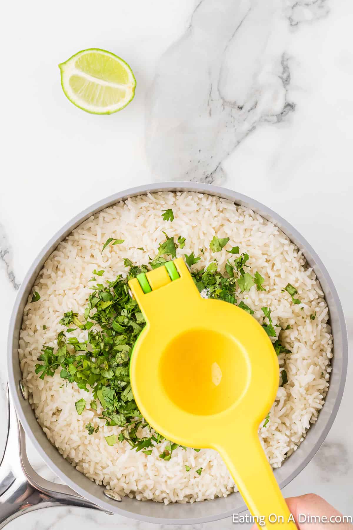 Overhead view of a bowl containing white rice garnished with chopped cilantro. A yellow citrus squeezer is placed on top of the rice, reminiscent of a Chipotle Cilantro Lime Rice Recipe. A halved lime and a silver serving spoon are placed nearby on a white marble surface.