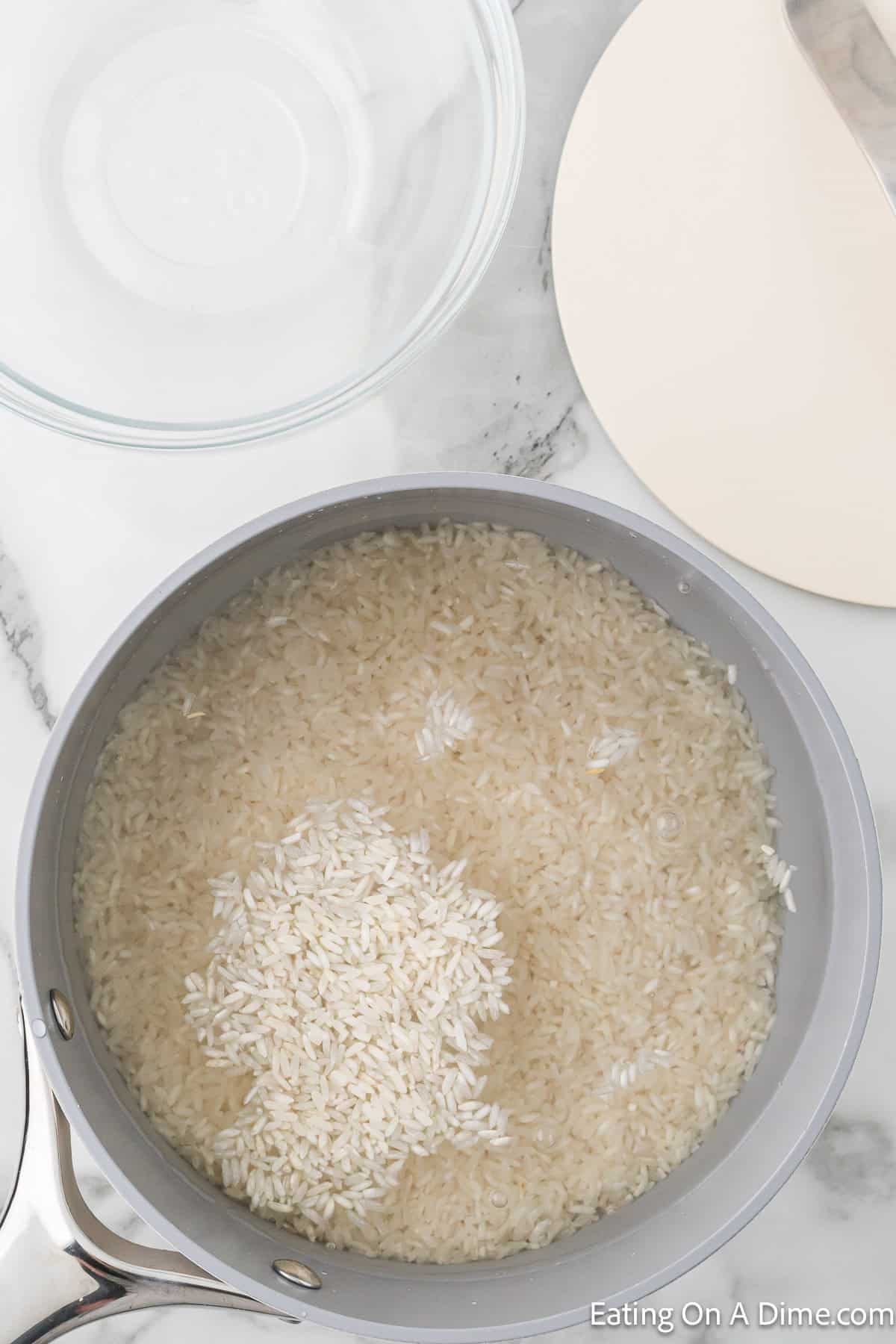 A cooking pot halfway filled with uncooked rice soaking in water is set on a marble countertop, ready to become part of the Chipotle Cilantro Lime Rice Recipe. Nearby, there's an empty clear glass bowl and a beige plate, along with a part of a metal pot handle visible in the lower-left corner.