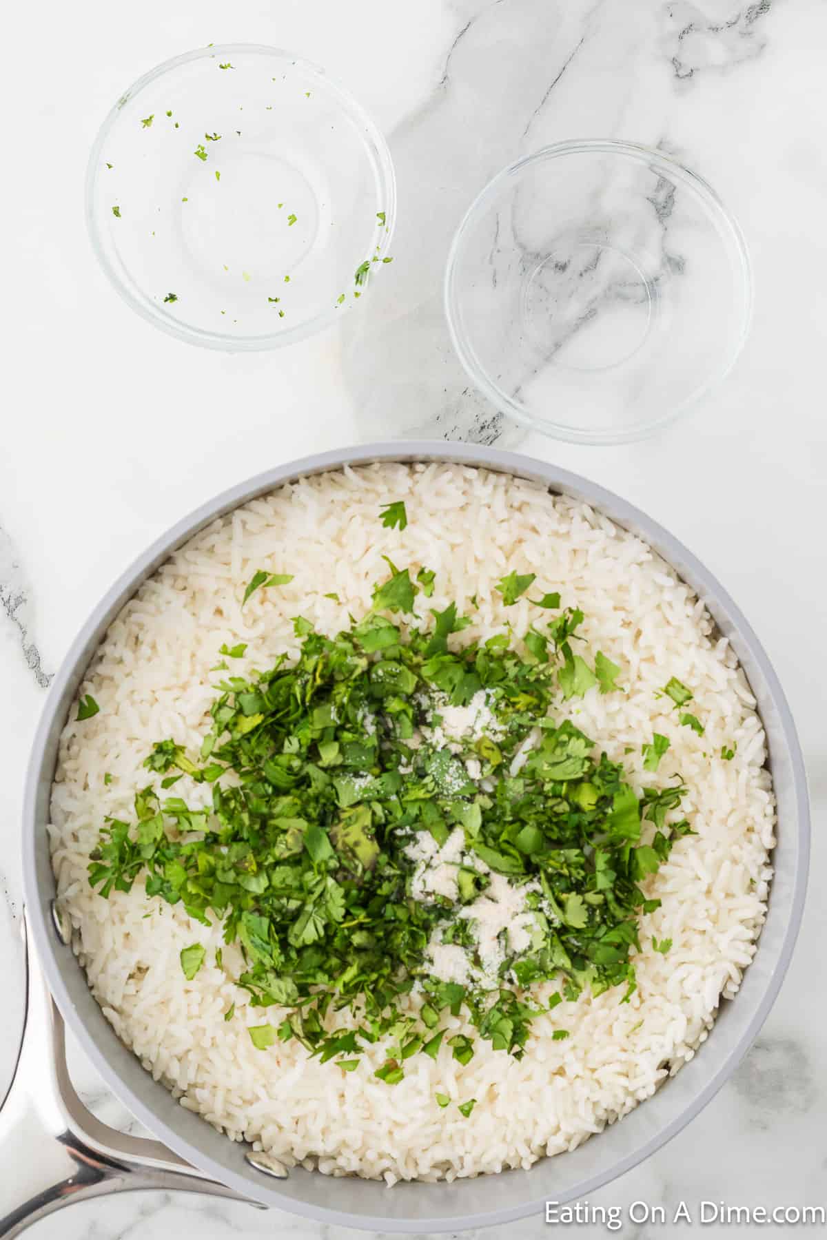 A pot of cooked rice is topped with finely chopped parsley and seasonings, resembling a Chipotle Cilantro Lime Rice Recipe. The pot is placed on a marble surface with two small empty glass bowls nearby. The image has a watermark "Eating On A Dime.com" in the bottom right corner.