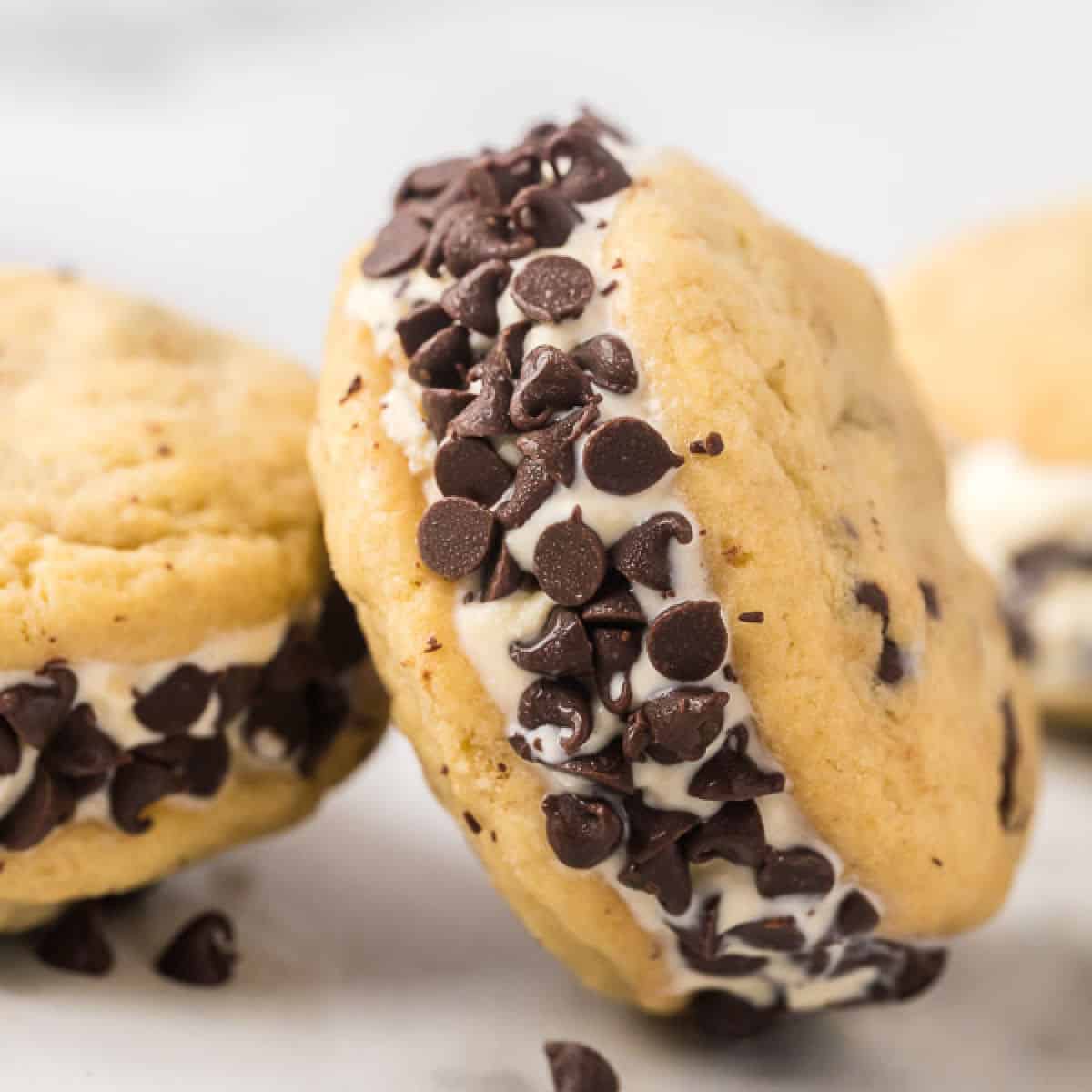 A close-up of a chocolate chip cookie ice cream sandwich with vanilla ice cream nestled between two soft and chewy cookies. The edges of the ice cream are coated in a layer of mini chocolate chips. Another sandwich peeks out enticingly from the background.