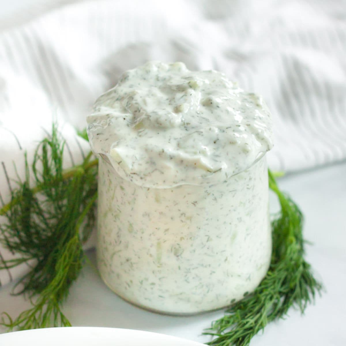 A glass jar filled with creamy ranch dressing, speckled with herbs, sits on a white surface. Fresh dill sprigs are arranged around the jar like in a tzatziki sauce recipe, with a white, textured cloth in the background.