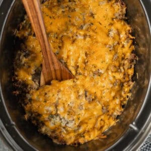 A cheesy crockpot casserole with ground meat and herbs in a slow cooker. A wooden spoon is partially submerged in the dish, which is topped with melted cheese, hash browns, and specks of green herbs, making for a delightful hamburger hashbrown casserole recipe.