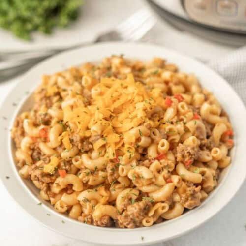 An Instant Pot Hamburger Helper recipe featuring creamy macaroni pasta mixed with ground beef and garnished with shredded cheese. Chopped herbs and red bell peppers add a pop of color atop this delightful dish, served on a white plate, with a fork and checkered napkin in the background.