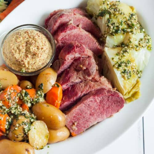 A plate of sliced corned beef, cooked cabbage with herb topping, small potatoes, and carrots. Prepared using an instant pot, this dish exudes comfort and flavor. A small glass bowl of whole grain mustard sits on the side, all arranged on a white oval plate.