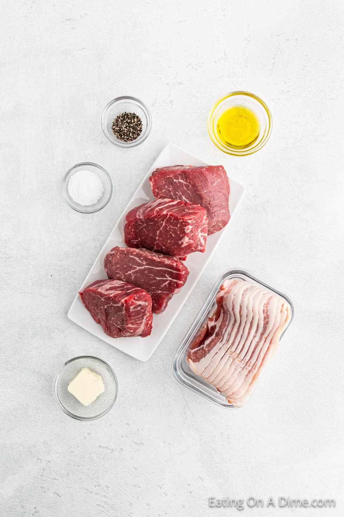 Slices of bacon sit in a glass container, ready to transform raw steak cuts into exquisite bacon-wrapped filet mignon. Accompanied by small bowls of olive oil, black pepper, salt, and a pat of butter, everything is elegantly arranged on a light gray surface.