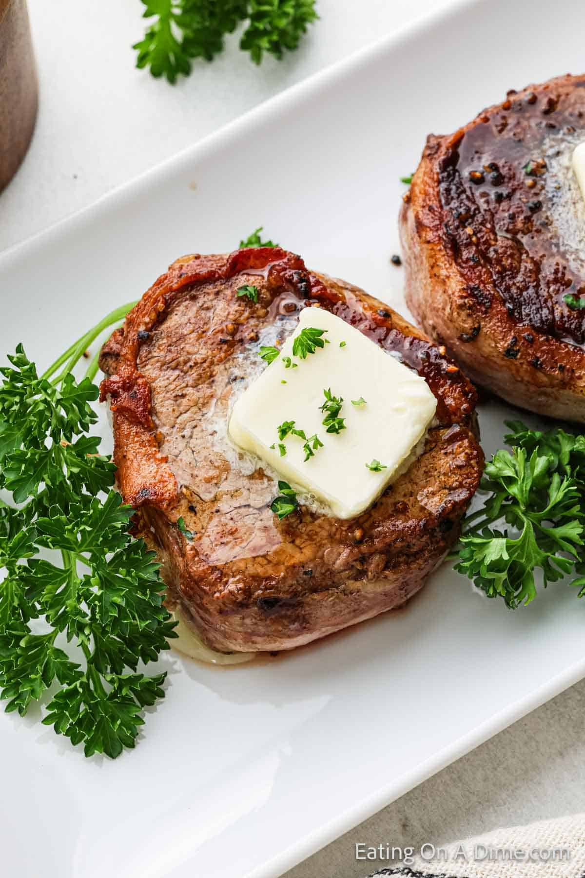 A close-up of a succulent bacon-wrapped filet mignon topped with a melting pat of butter and garnished with chopped parsley. It is served on a white plate with sprigs of fresh parsley on the side.