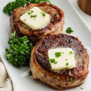 Two grilled steaks, reminiscent of bacon-wrapped filet mignon, topped with melting butter and garnished with parsley on a white plate. Fresh parsley sprigs are placed beside the steaks.