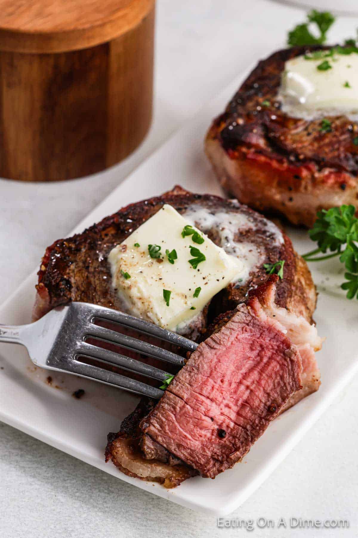 A juicy, medium-rare bacon-wrapped filet mignon topped with a melted pat of butter and garnished with chopped herbs on a white plate. A fork holds a cut piece of the steak. Another steak and a wooden container are in the background.