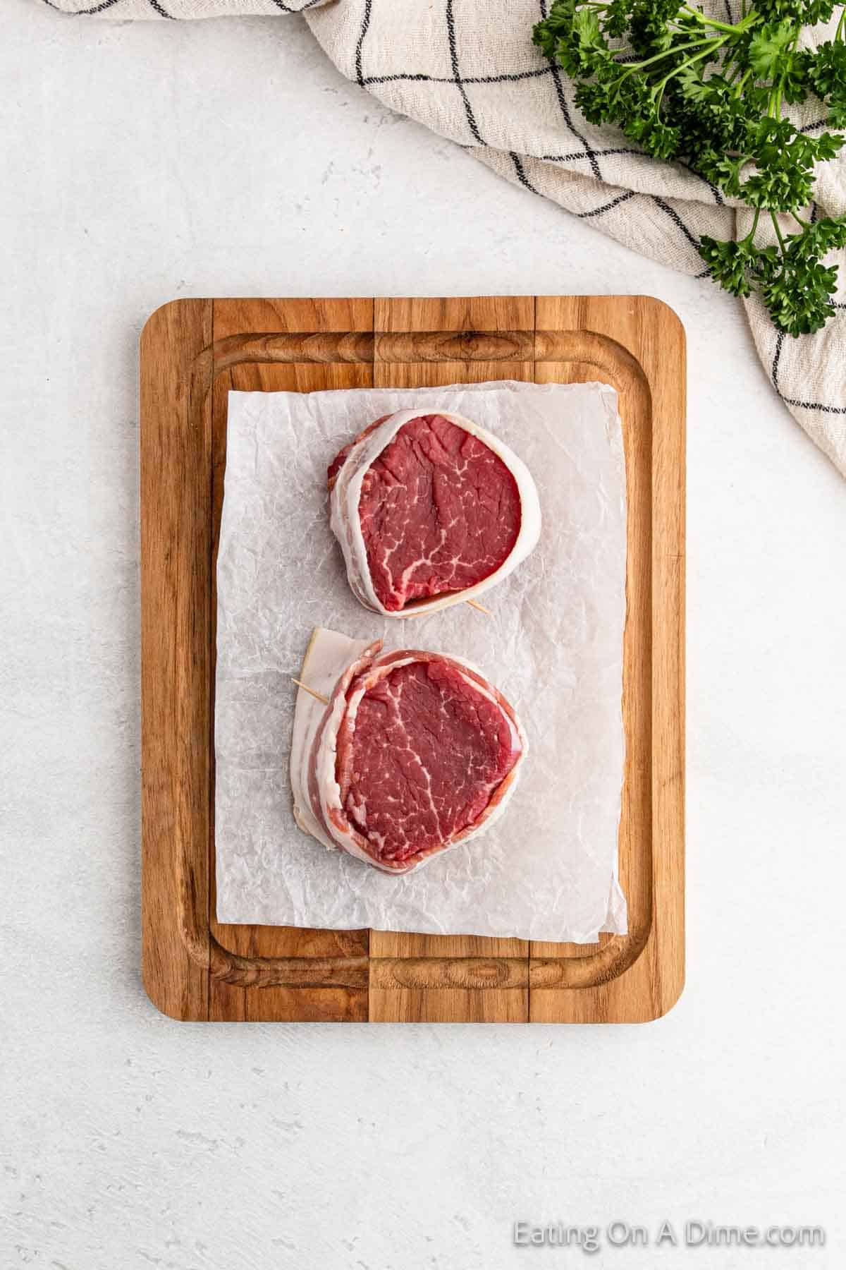 Two succulent bacon-wrapped filet mignon steaks rest on parchment paper atop a wooden cutting board. A striped cloth and fresh parsley decorate the upper corners on a light gray surface, enhancing the gourmet presentation of these premium cuts.