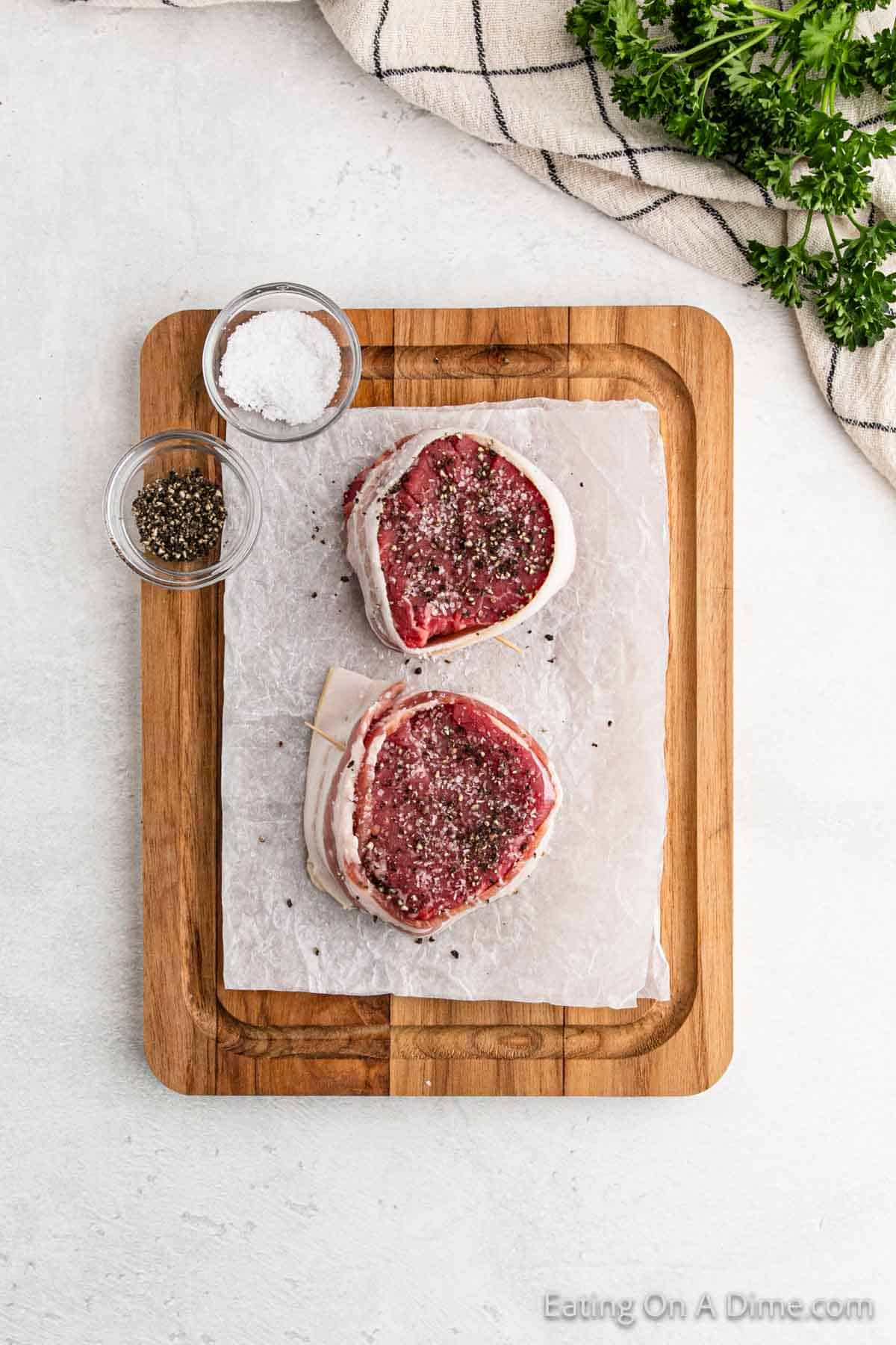 Two raw steaks, soon to become succulent bacon-wrapped filet mignon, are seasoned with black pepper on a parchment-lined wooden cutting board. Nearby are small bowls of salt and pepper. Fresh herbs and a striped cloth enhance the inviting culinary scene.