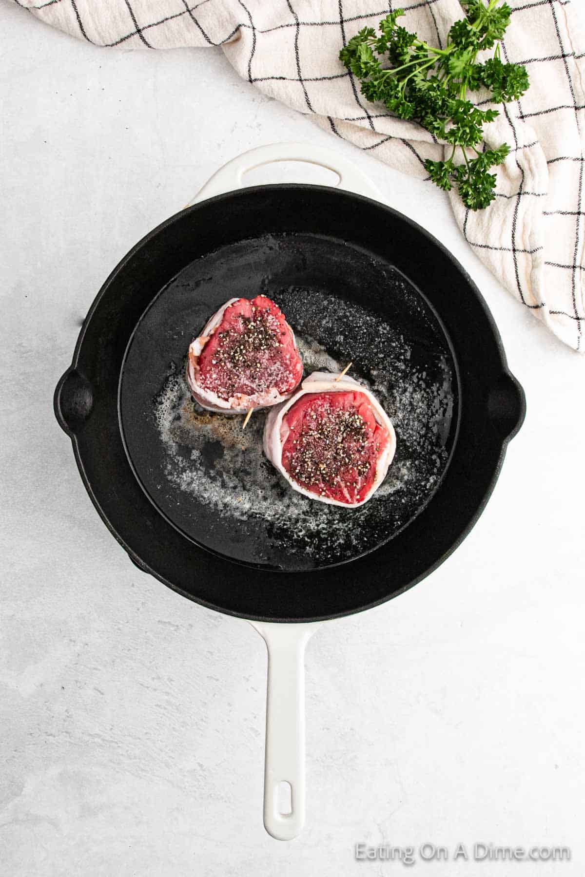 Two bacon-wrapped filet mignons sizzle in a black skillet, surrounded by melted butter. Beside the skillet, there's a bunch of fresh parsley and a striped kitchen towel resting on a light gray surface.